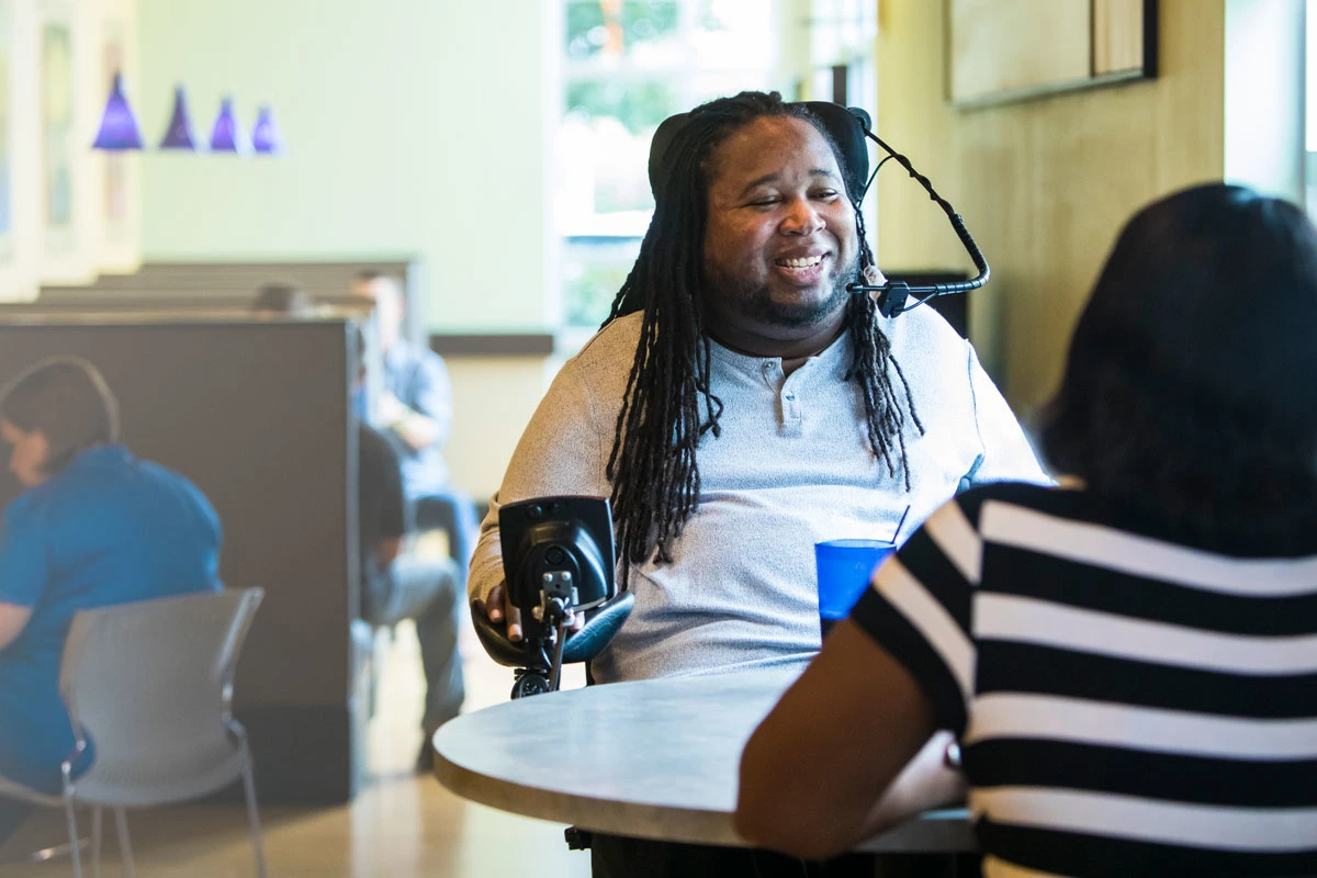 Eric sits at a table, smiling and talking to his friend